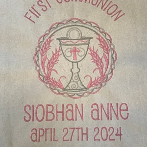 May include: A beige fabric with pink embroidery. The embroidery features a chalice with a cross, wheat stalks, and the words "First Communion". Below the design is the name "Siobhan Anne" and the date "April 27th 2024".