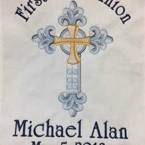 May include: White embroidered fabric with a blue cross and the text "First Communion" above and "Michael Alan May 5, 2018" below.