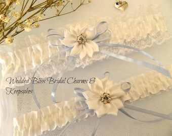 May include: Two white satin garters with blue ribbon accents and a white flower with a rhinestone center. The garters are on a white background with a small charm that says "Something Blue".