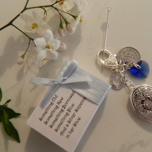 May include: A small white box with a blue ribbon tied around it. The box has text that reads "Something Old, Something New, Something Blue, Something Borrowed, find a Silver Sixpence in her Shoe".  A silver charm with a locket, horseshoe, heart, and blue crystal hangs from a silver chain.