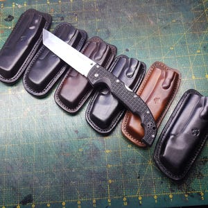 Puede incluir: Seis fundas de cuero para cuchillos en negro y marrón, con un cuchillo negro y plateado con mango texturizado.