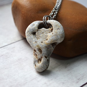 Könnte beinhalten: Eine einzigartige Anhänger Halskette mit einem natürlichen, hellgrauen Stein mit einer ausgeprägten Form und Textur. Der Stein hängt an einer silberfarbenen Kette. Der Anhänger hat ein Loch in der Nähe der Oberseite und wird auf einem braunen Stein präsentiert.