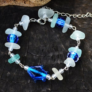 Könnte beinhalten: Ein Armband mit einer silberfarbenen Metallkette, durchsetzt mit mattem weißem und hellgrünem Meerglas, klaren Glasperlen, facettierten königsblauen Glasperlen und einem einzelnen blauen Glasanhänger in Form eines Delfins mit Karabinerverschluss.