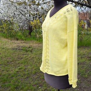 Puede incluir: Un suéter amarillo pálido de manga larga con cuello en V y delicados detalles calados a lo largo del escote y las mangas. El suéter se exhibe en un maniquí, mostrando su tejido ligero y su ajuste relajado.