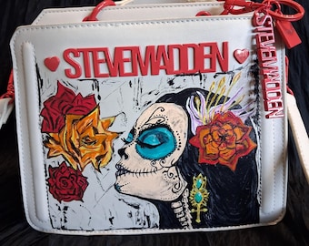 Peut inclure: Un sac à main blanc structuré avec le texte rouge STEVE MADDEN et une illustration graphique d'une personne au maquillage de tête de mort, entourée de roses rouges et orange. Une bandoulière à chaîne rouge et une étiquette rouge sont fixées.