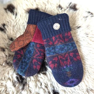 Puede incluir: Un par de mitones hechos a mano con pu&ntilde;os acanalados azul marino y un bot&oacute;n decorativo. Los mitones presentan un colorido patr&oacute;n geom&eacute;trico en tonos rojos, azules y naranjas. Est&aacute;n hechos de un material suave y c&aacute;lido.