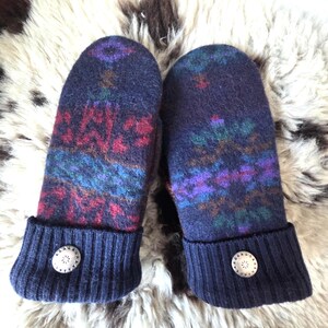 Puede incluir: Un par de mitones de lana azul marino con un colorido estampado geom&eacute;trico en rojo, morado, verde y azul. Los pu&ntilde;os son de punto acanalado azul marino con un bot&oacute;n decorativo. Los mitones se exhiben sobre una superficie clara y texturizada.