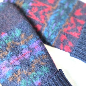 Puede incluir: Un par de mitones de lana azul marino con un colorido patr&oacute;n geom&eacute;trico en rojo, morado, verde y azul. Los mitones tienen un pu&ntilde;o acanalado y un peque&ntilde;o bot&oacute;n redondo.