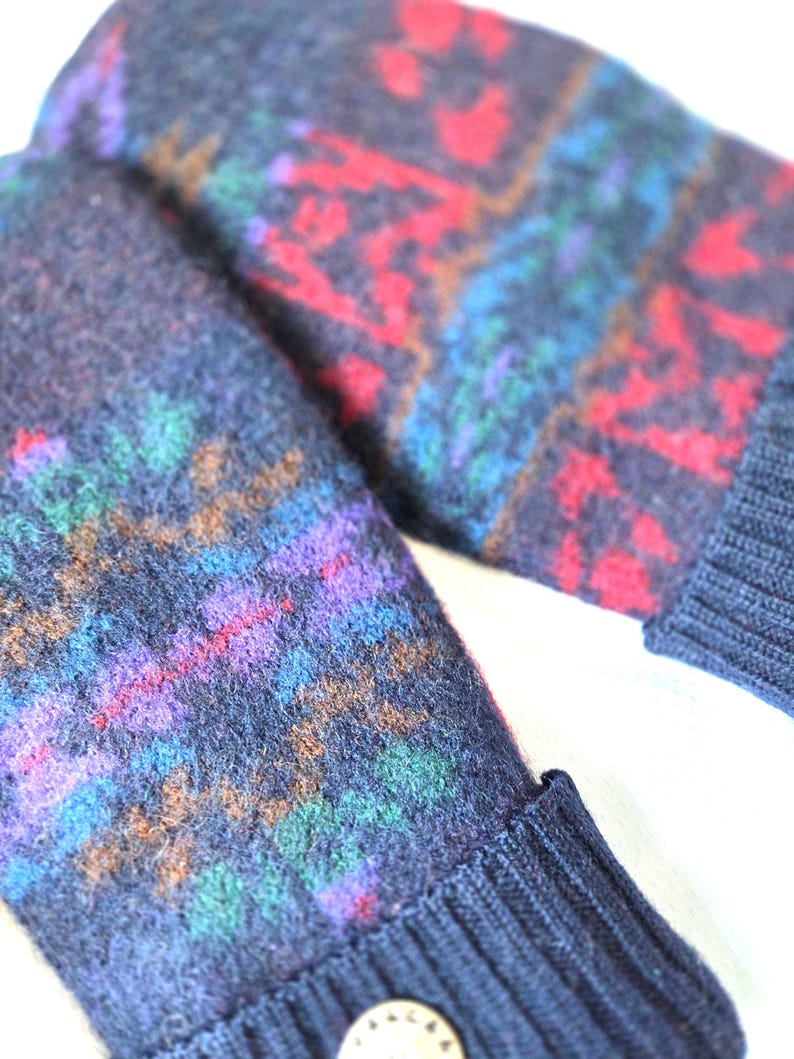 Puede incluir: Un par de mitones de lana azul marino con un colorido patr&oacute;n geom&eacute;trico en rojo, morado, verde y azul. Los mitones tienen un pu&ntilde;o acanalado y un peque&ntilde;o bot&oacute;n redondo.