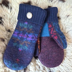 Puede incluir: Un par de mitones hechos a mano en tonos de azul, morado y burdeos. Los mitones presentan un dise&ntilde;o estampado en la parte superior y un color liso en la palma. Un bot&oacute;n de madera adorna el pu&ntilde;o de un mit&oacute;n. Los mitones est&aacute;n hechos de un material suave y texturizado.