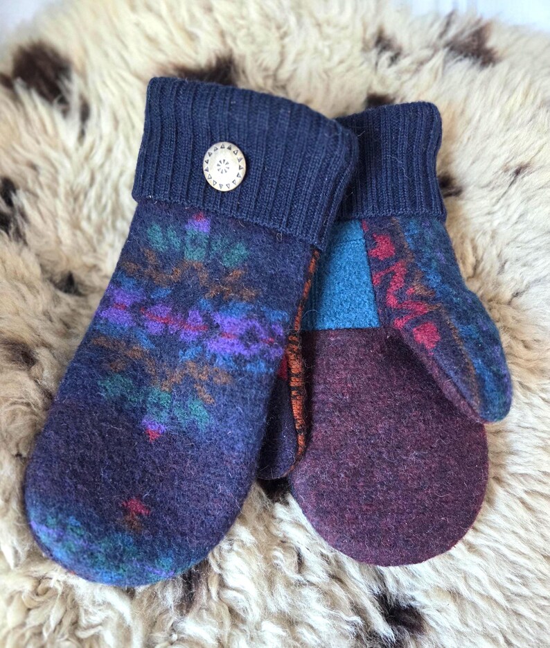 Puede incluir: Un par de mitones hechos a mano en tonos de azul, morado y burdeos. Los mitones presentan un dise&ntilde;o estampado en la parte superior y un color liso en la palma. Un bot&oacute;n de madera adorna el pu&ntilde;o de un mit&oacute;n. Los mitones est&aacute;n hechos de un material suave y texturizado.