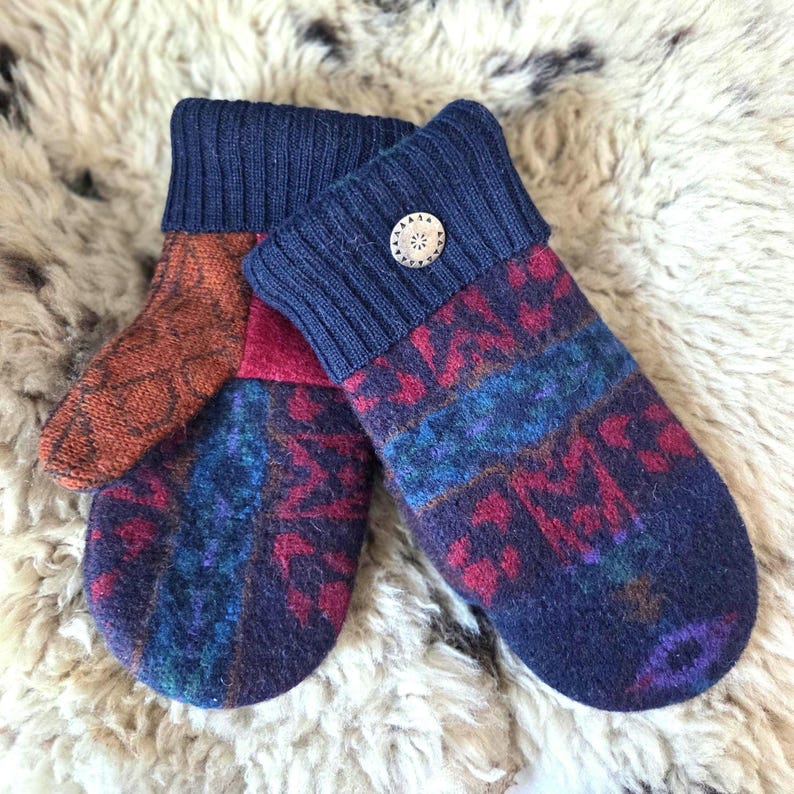 Puede incluir: Un par de mitones hechos a mano con pu&ntilde;os acanalados azul marino y un bot&oacute;n decorativo plateado. Los mitones presentan un patr&oacute;n colorido en tonos azules, rojos y naranjas. Est&aacute;n hechos de un material suave y texturizado.