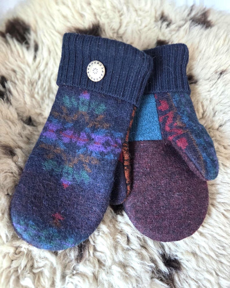 Puede incluir: Un par de mitones hechos a mano en tonos azul marino, morado y verde azulado. Los mitones presentan un dise&ntilde;o estampado en la parte delantera y un color morado liso en las palmas. Los pu&ntilde;os son de color azul marino con un bot&oacute;n decorativo.
