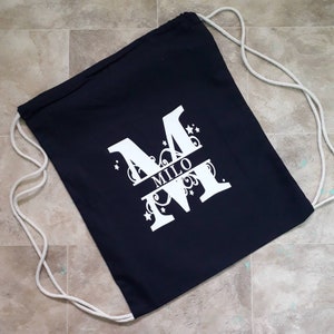 Puede incluir: Una bolsa con cordón de color azul marino con una gran letra "M" blanca y el nombre "MILO". El diseño incluye remolinos y estrellas. La bolsa tiene cordones blancos. Un accesorio práctico y elegante.