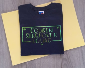 May include: A navy blue t-shirt with a green and gold foil design that says "Cousin Sleepover Squad" inside a rectangle with stars on either side.