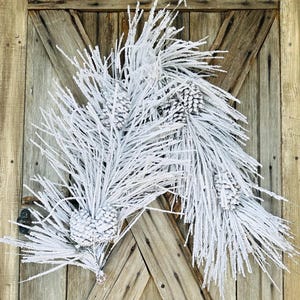 May include: A white frosted pinecone and branch wreath. The wreath is composed of white frosted pine needles and several pinecones. The wreath is mounted on a rustic wooden door with a diagonal cross design.