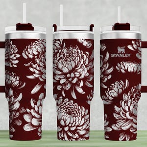 May include: A maroon stainless steel tumbler with a white floral design and a black handle. The tumbler has a lid with a straw hole and the word "STANLEY" is printed on the side.