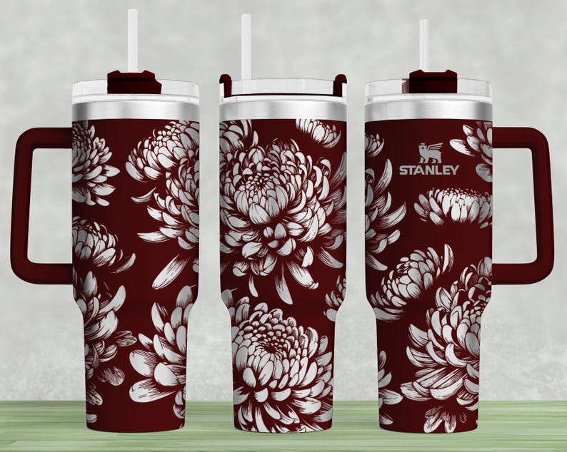 May include: A maroon stainless steel tumbler with a white floral design and a black handle. The tumbler has a lid with a straw hole and the word "STANLEY" is printed on the side.