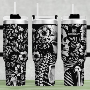 May include: Black and white stainless steel tumbler with a floral and skeletal design. The tumbler has a handle and a straw lid. The text "STANLEY" is printed on the tumbler.
