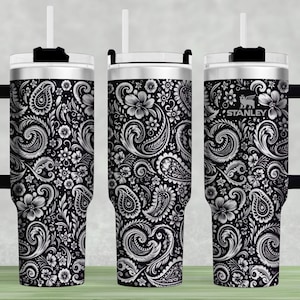 May include: A set of three Stanley tumblers with a black and white paisley floral design. Each tumbler has a black handle, a white lid, and a white straw. The Stanley logo is visible on one of the tumblers.