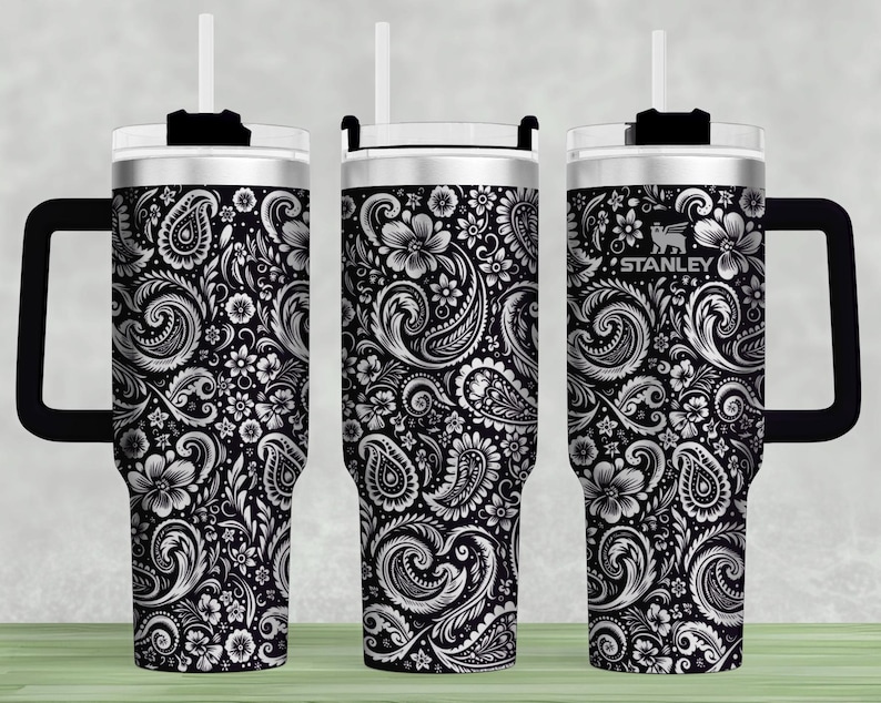 May include: A set of three Stanley tumblers with a black and white paisley floral design. Each tumbler has a black handle, a white lid, and a white straw. The Stanley logo is visible on one of the tumblers.