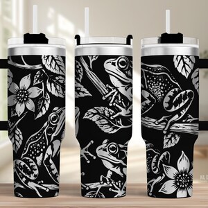 Puede incluir: Un vaso de acero inoxidable en blanco y negro con asa, tapa transparente y pajita. El exterior presenta un patrón gráfico repetitivo de ranas posadas en ramas rodeadas de flores y hojas detalladas. El texto KL Design es visible.