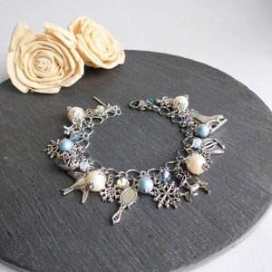 Peut inclure: Un bracelet à breloques en argent avec une variété de breloques, dont un flocon de neige, un oiseau, un miroir et un patin. Les breloques sont attachées à une chaîne en argent avec des perles blanches et bleues.