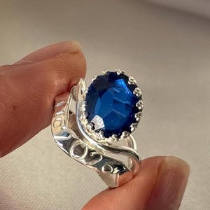 May include: A silver ring with a large, oval-cut, deep blue gemstone. The gemstone is set in a crown-like bezel. The ring's band has an ornate, swirling design.