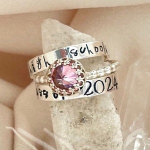 May include: A stack of three silver rings. The top ring says "high school" in a handwritten font. The middle ring features a pink gemstone with a decorative setting. The bottom ring has the year "2024" and the numbers "199".