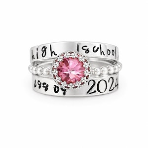 Könnte beinhalten: Ein Satz von drei silbernen Ringen. Der obere Ring trägt die Aufschrift "high school", der mittlere Ring hat ein Perlendesign und der untere Ring trägt die Aufschrift "199 of 2024". Der zentrale Ring hat einen rosa Edelstein mit dekorativem Rand.