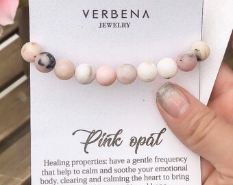 Könnte beinhalten: Rosa Opal-Armband mit weißen und rosafarbenen Perlen auf einer weißen Karte mit dem Text "VERBENA JEWELRY" und "Pink opal". Die Karte enthält auch den Text "Heileigenschaften: haben eine sanfte Frequenz, die helfen, Ihren emotionalen Körper zu beruhigen und zu besänftigen, das Herz zu reinigen und zu beruhigen, um ein Gefühl von Liebe, Frieden und Hoffnung zu bringen."