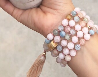 May include: A stack of beaded bracelets featuring pink, blue, and yellow gemstones. The bracelets are strung on pink cord and accented with a gold-colored square bead and a peach-colored tassel.
