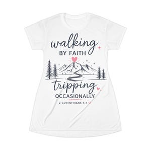 Puede incluir: Una camiseta blanca de manga corta con un gráfico de montañas, pinos y un camino sinuoso. El texto dice "walking BY FAITH tripping OCCASIONALLY" con un corazón rosa sobre las montañas y "2 CORINTHIANS 5:7" en la parte inferior.