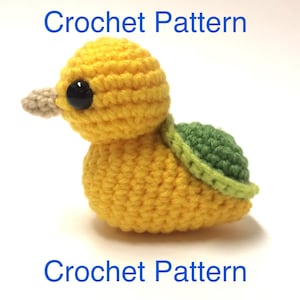 Può includere: Schema all'uncinetto per un'anatra gialla con un guscio verde. L'anatra ha occhi neri e un becco beige.