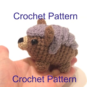 May include: Crochet pattern for a small brown bear wearing a purple hat. The bear is sitting on a hand.
