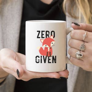 May include: White ceramic mug featuring a cartoon fox in shades of red and cream. The mug has the words "ZERO GIVEN" in black block letters. The mug is held by a person.