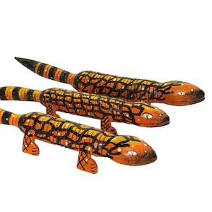 May include: Three hand-painted lizard figurines arranged in a row. Each lizard features a vibrant orange base color with a distinct black net-like pattern across the body and tail. The tails are striped with black and orange, and the heads have eyes.