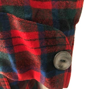 May include: A long-sleeved garment cuff featuring a vibrant red, dark blue, and forest green plaid pattern. The cuff is secured with a single, round, dark grey button that is centered on the rounded tab of the sleeve fabric.
