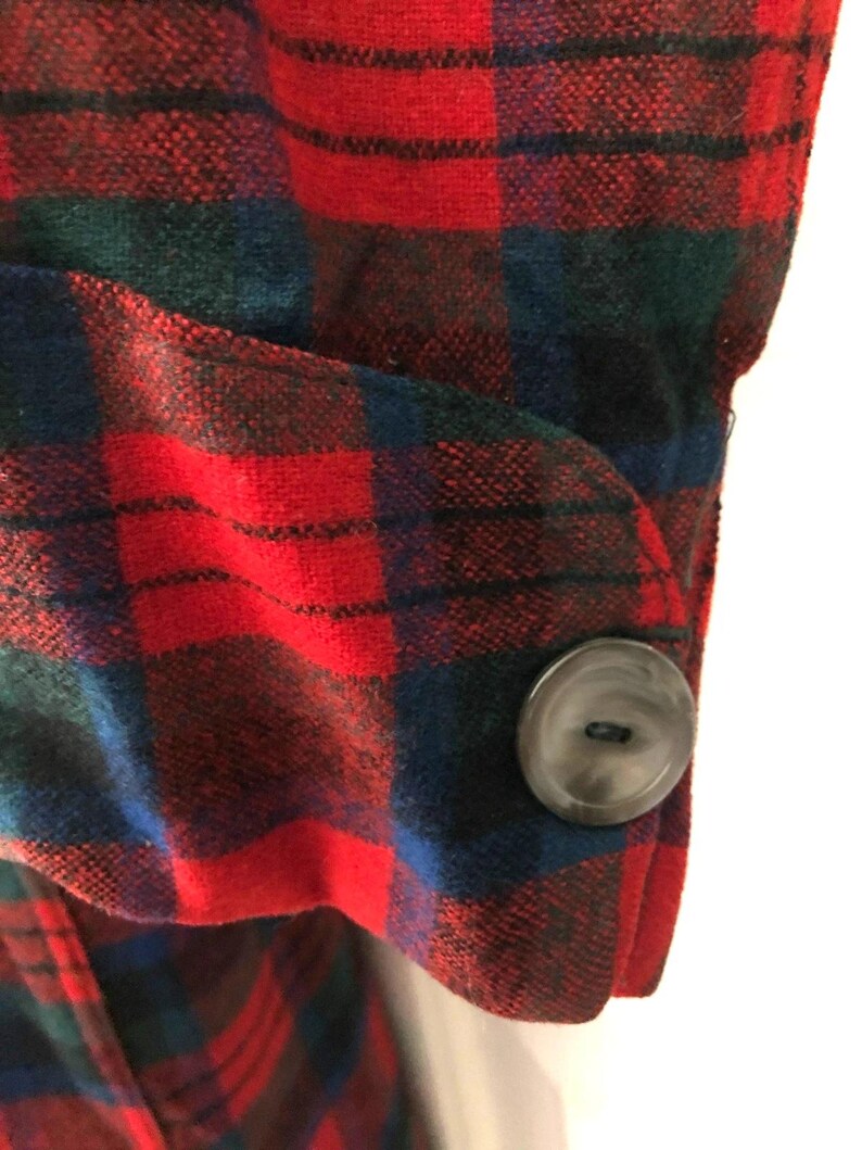 May include: A long-sleeved garment cuff featuring a vibrant red, dark blue, and forest green plaid pattern. The cuff is secured with a single, round, dark grey button that is centered on the rounded tab of the sleeve fabric.
