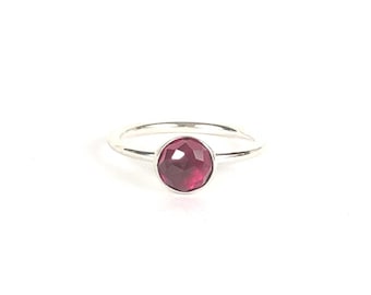 Könnte beinhalten: Ein silberner Ring mit einem facettierten, runden, tiefroten Edelstein. Der Edelstein ist in einem schlichten, polierten Silberband gefasst. Der Ring wird vor einem weißen Hintergrund gezeigt, um sein elegantes Design hervorzuheben.