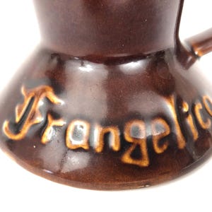 May include: Close-up of a brown ceramic coffee pot with a handle. The word "Frangelico" is written in a gold colour on the base of the pot. The pot has a glossy finish and a unique shape.