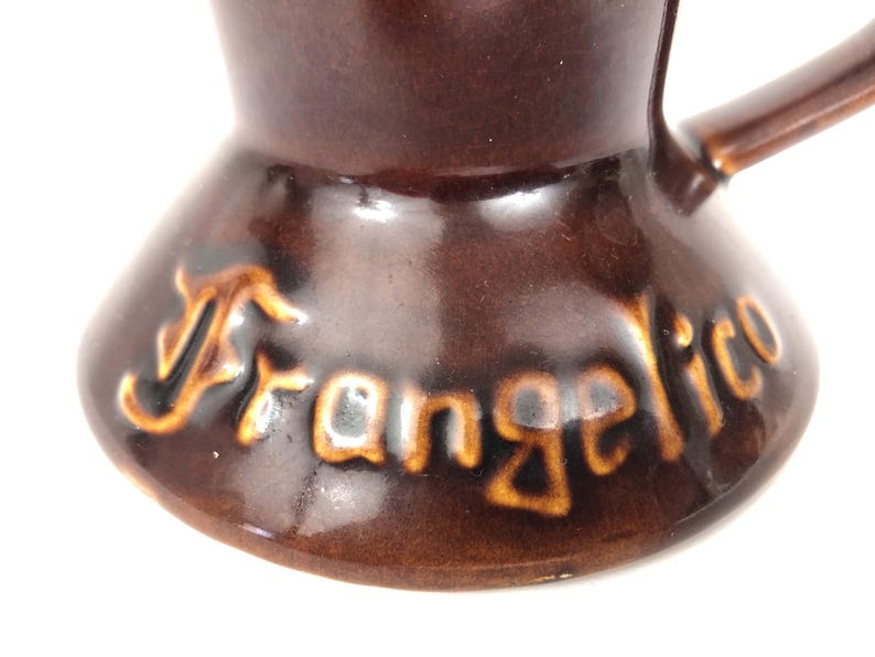 May include: Close-up of a brown ceramic coffee pot with a handle. The word "Frangelico" is written in a gold colour on the base of the pot. The pot has a glossy finish and a unique shape.