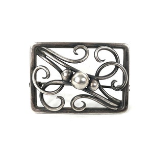 May include: A silver brooch with a rectangular frame and a delicate, swirling design. The design features three round, silver beads.