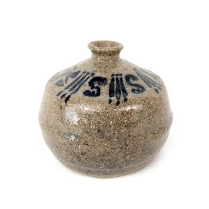 May include: A handmade, speckled ceramic vase with a rounded body and a narrow neck. The vase is a neutral tan colour with dark blue abstract designs and the letters "S" and "I".