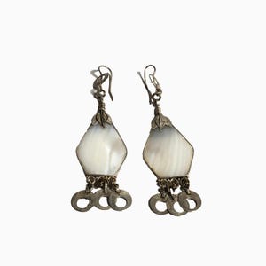 May include: A pair of silver-toned drop earrings featuring a central diamond-shaped white shell inlay. Each earring is topped with a small metal leaf detail and finished at the bottom with three dangling circular metal rings attached by small links.