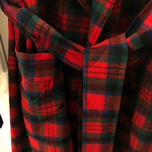May include: A robe featuring a red, dark green, and blue plaid pattern. The garment includes a matching fabric belt tied at the waist and a visible patch pocket on the left side. The fabric has a soft, textured appearance with a classic tartan design.