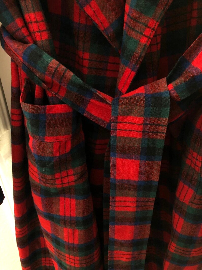May include: A robe featuring a red, dark green, and blue plaid pattern. The garment includes a matching fabric belt tied at the waist and a visible patch pocket on the left side. The fabric has a soft, textured appearance with a classic tartan design.
