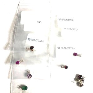 May include: A collection of small gemstone pendants in individual clear plastic bags with printed labels. The pendants feature various colored stones including red, purple, and green, each set in a metal prong setting with a small bail for hanging.