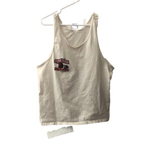 May include: Cream-colored tank top with a scoop neck, hanging on a white hanger. The front features a graphic with the text "Royal Flush" in red and black, with additional text below. The tank top is made of a soft, lightweight material.