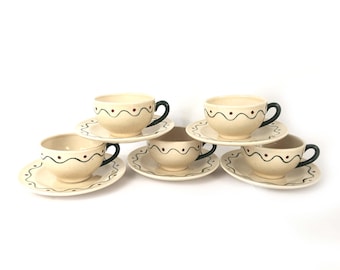 May include: A collection of six cream-coloured ceramic teacups and saucers. Each cup features a dark green handle and is decorated with a wavy black line and small red dots. The saucers have a matching design.